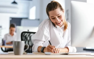 Die Top Funktionen für einen B2B-Online Shop - Frau sitzt telefonierend am PC und notiert sich Tipps mit einem Stift